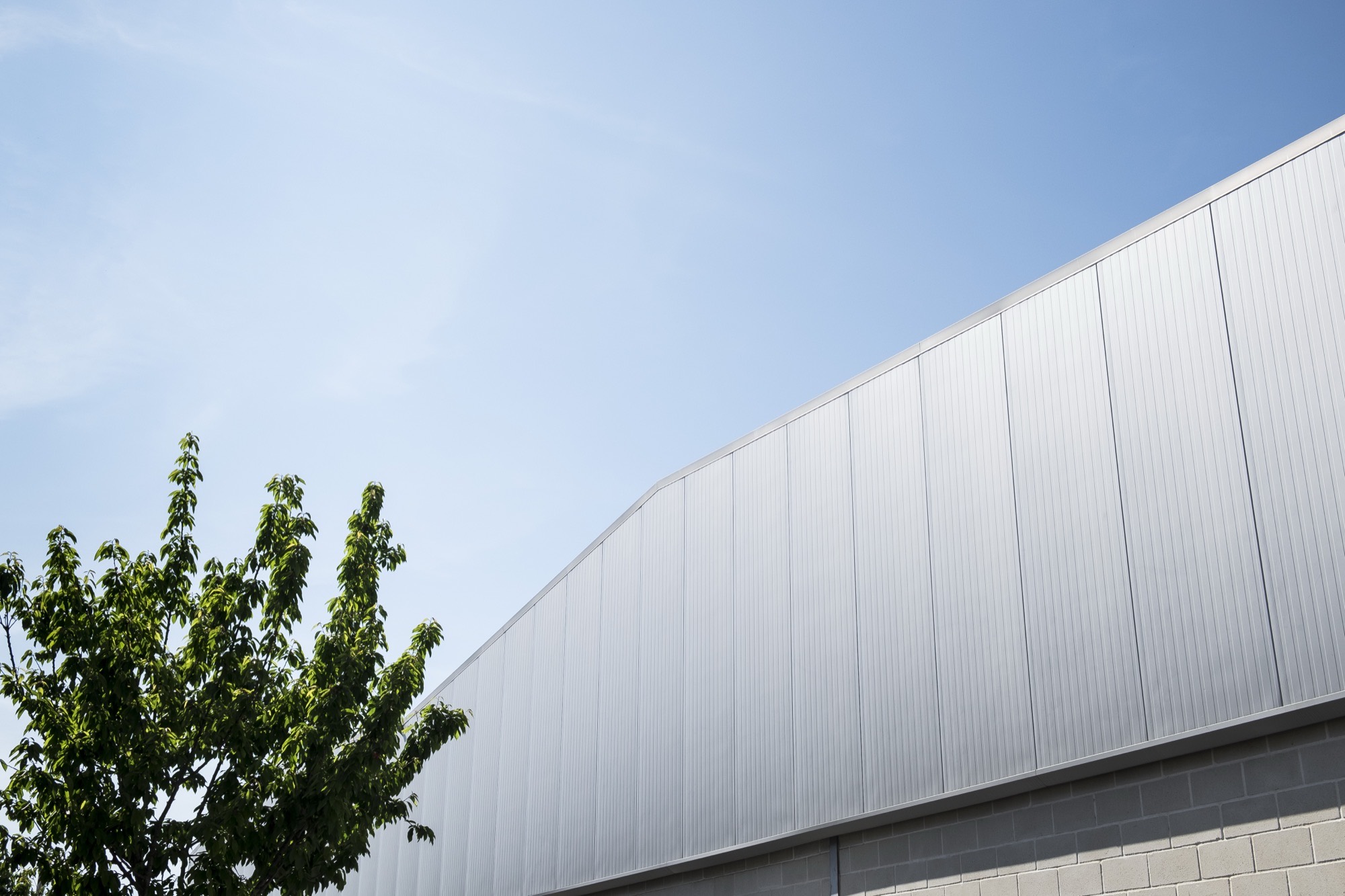 Fachada exterior do pavilhão da JVperfis: parede de chapa perfilada prateada a subir na diagonal contra um céu azul limpo, com uma árvore verde no canto inferior esquerdo e embasamento em blocos de cimento.