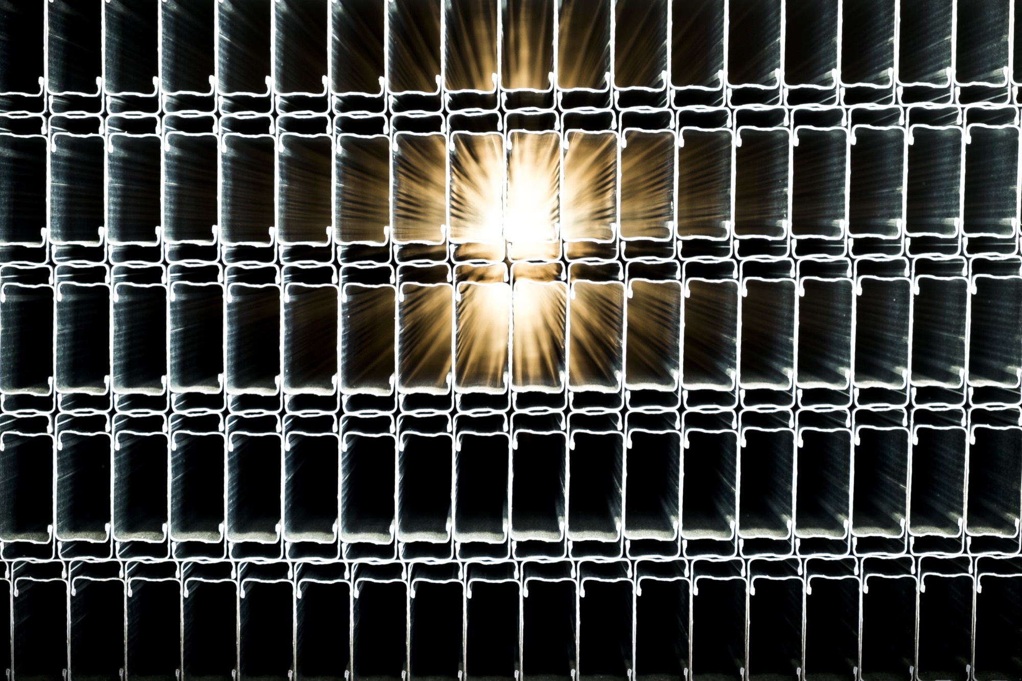 Grelha retroiluminada de secções transversais de perfis montante empilhados, com o sol a irromper em estrela através de duas aberturas na grelha metálica escura.